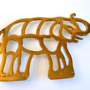 Może przedstawiać: Złota, ażurowa figurka słonia. Słoń przedstawiony jest w stylizowanej, uproszczonej formie z dużymi uszami, trąbą i czterema nogami. Metal ma lekko postarzany wygląd, co sugeruje, że może być w stylu vintage lub antycznym.