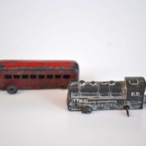 Peut inclure: Deux jouets anciens : un bus rouge et un train noir. Le bus est rectangulaire avec de petites fenêtres et un aspect usé. Le train a un moteur détaillé et une goupille saillante. Les deux jouets sont sur fond blanc.