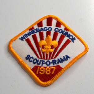 May include: A diamond-shaped embroidered patch with a yellow border. The design features a white background with red sunburst rays, a yellow fleur-de-lis, and blue text reading Winnebago Council Scout-O-Rama 1987 in yellow and blue thread.