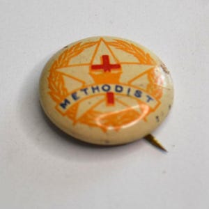 May include: A vintage, circular pin badge with a cream-coloured background. The badge features a star and cross design in orange and red, with the word "METHODIST" in orange lettering. A gold pin extends from the back.