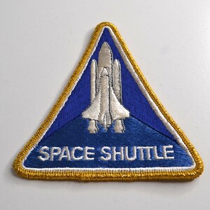 Può includere: Una toppa ricamata triangolare che raffigura una navetta spaziale bianca centrata su uno sfondo blu. La toppa è bordata da un filo dorato spesso e da un sottile bordo interno bianco. Il testo SPACE SHUTTLE è ricamato in bianco.