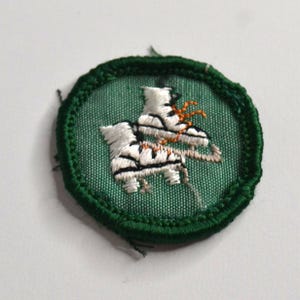 May include: A circular embroidered patch featuring a green fabric background with a dark green stitched border. The center displays a white pair of ice skates with black blade details and orange laces, rendered in detailed thread embroidery.