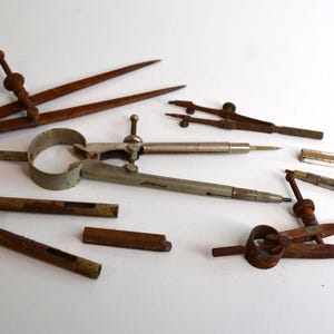 May include: A collection of vintage drafting tools including four metal compasses with varying degrees of rust and oxidation, a small metal lead holder, and a separate metal component, all arranged together on a plain white surface.