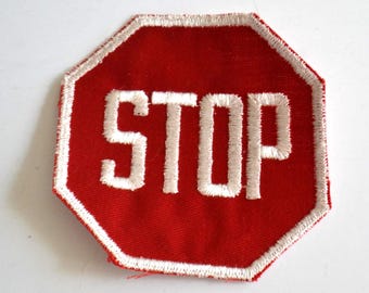 Könnte beinhalten: Roter achteckiger Aufnäher mit dem Wort „STOP“ in weißen Großbuchstaben. Der Aufnäher hat einen weiß gestickten Rand. Der Aufnäher ist wahrscheinlich ein Applikations- oder Bügelbild.