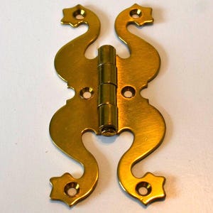 Puede incluir: Una bisagra decorativa de latón con un diseño único y estilizado. La bisagra presenta un barril central y dos hojas ornamentadas en forma de S. Cada hoja tiene un extremo en forma de estrella con un agujero para el montaje. El latón tiene un tono dorado cálido.