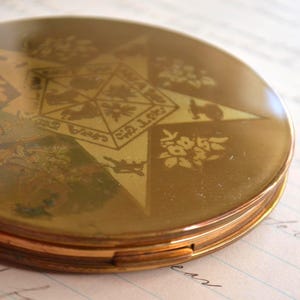 May include: A vintage gold-toned compact with a starburst design. The compact features floral and geometric patterns, with a central star shape. The compact is round and has a hinged opening, with a gold rim.