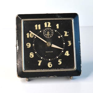 May include: A vintage black Westclox alarm clock with a square face. The clock has gold-colored numerals and hands, with a smaller dial at the top. The text "WESTCLOX" and "MADE BY WESTCLOX LASALLE ILL USA" are visible.