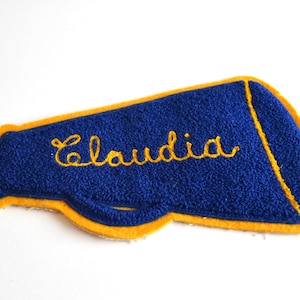 Könnte beinhalten: Blau-goldener Cheerleader-Megaphon-Aufnäher mit dem Namen "Claudia" in goldener Schrift gestickt. Der Aufnäher hat ein strukturiertes, filzartiges Aussehen und einen gelben Rand.