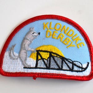 Pode incluir: Patch bordado com borda vermelha. Apresenta um fundo azul claro com uma figura de cão branco, um sol amarelo e um trenó preto. O texto "KLONDIKE DERBY" é amarelo.