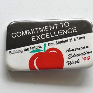 May include: A rectangular pinback button featuring a black top section with the white text COMMITMENT TO EXCELLENCE and a white bottom section with the text Building the Future, One Student at a Time, American Education Week '94 and a red apple.