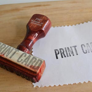 May include: A dark brown wooden rubber stamp with a handle featuring the text JVC ELKHART, IN 219-293-0113. The stamp base displays the text PRINT CARD, which is also clearly stamped in black ink on a piece of white paper with jagged edges.