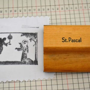 May include: A light-colored wooden rubber stamp with the text St. Pascal printed on the side. Next to the stamp is a white piece of paper with a black ink impression of an angel holding a monstrance toward a figure with a halo near a tree.