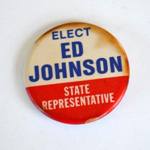 May include: A vintage campaign button with a cream-colored top and a red bottom. The button reads "ELECT ED JOHNSON STATE REPRESENTATIVE" in blue and white text. The button has a slightly aged appearance.