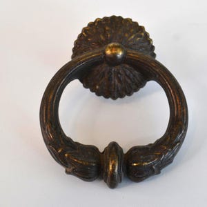 May include: An antique bronze door knocker with a circular handle and a decorative backplate. The handle is adorned with leaf-like details, and the backplate has a sunburst design. The knocker is made of metal and has a vintage aesthetic.