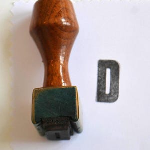 Könnte beinhalten: Ein Holzbuchstabenstempel mit einem dunkelbraunen Griff und einer rechteckigen Basis. Der Stempel befindet sich neben einem gedruckten Buchstaben "D". Die Basis des Stempels hat ein grünes Kissen. Der Buchstabe "D" ist in Dunkelgrau gedruckt.