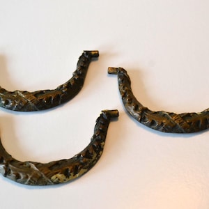 Op de afbeelding: Drie bronzen, gebogen handgrepen voor laden. Elke handgreep heeft een textuur, organisch ontwerp en een klein, rechthoekig eindstuk. De handgrepen zijn gerangschikt op een wit oppervlak.