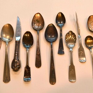 Puede incluir: Una colección de cucharas de plata vintage y un cuchillo para mantequilla. Las cucharas son de varios tamaños y estilos, incluyendo una cuchara con forma de concha y una cuchara pequeña con un mango decorativo. El cuchillo para mantequilla tiene un mango de madera.