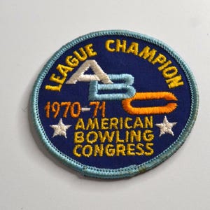 May include: A circular embroidered patch with a dark blue background and a light blue border. It features the yellow text LEAGUE CHAMPION, 1970-71, and AMERICAN BOWLING CONGRESS. The centre displays the letters ABC in white, blue, and orange.