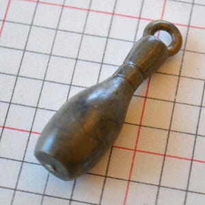 Puede incluir: Un amuleto de metal plateado en forma de pin de bolos con un bucle en la parte superior para sujetarlo a un collar o pulsera.
