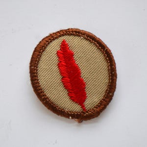 May include: A brown oval-shaped patch with a red embroidered feather in the center.