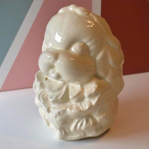 Könnte beinhalten: Weiße Keramikfigur eines Hundes mit geneigtem Kopf. Der Hund hat einen flauschigen Kragen und sitzt auf einem Sockel mit Blumenmuster.