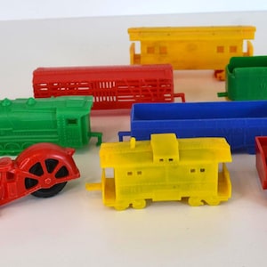 May include: A collection of colorful toy vehicles, including a red motorcycle, a green train engine, and various train cars in red, yellow, blue, and green. The toys are made of plastic and are arranged on a white surface.