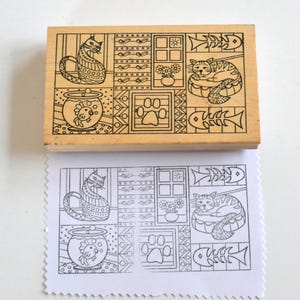 May include: A rectangular wooden stamp featuring a black and white design of cats, fish, paw prints, and floral elements. The stamp is pressed onto a white paper, revealing the stamped image. The design is in a block style.
