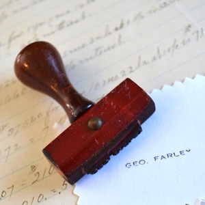 Może przedstawiać: Zabytkowy drewniany stempel z ciemnobrągową rączką i czerwoną prostokątną główką. Stempel jest odbity na białej kartce z czarnym napisem "GEO. FARLEY". Tło zawiera odręczne notatki.