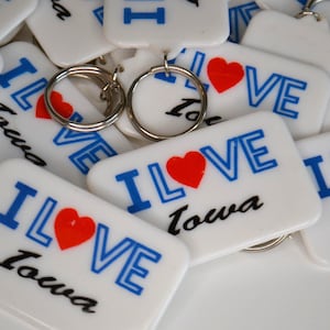 May include: White rectangular keyrings with silver key rings. The keyrings have the words "I LOVE Iowa" printed in blue and black, with a red heart replacing the word "love".