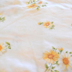 Puede incluir: Tela blanca con un estampado de margaritas amarillas y naranjas con hojas verdes. Las flores están dispersas por la tela, creando un diseño delicado y natural. La tela tiene una textura suave y ligeramente arrugada.