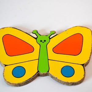 Peut inclure: Un morceau de puzzle en bois en forme de papillon. Le papillon a des ailes jaunes avec des accents orange et bleus. Le corps est vert avec un visage souriant. Les bords du bois sont visibles.