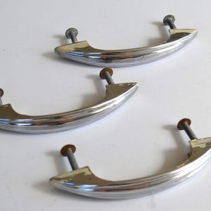 Pode incluir: Três puxadores de armário cromados vintage com um design curvo e canelado. Cada puxador tem um parafuso para montagem. Os puxadores são prateados com algum desgaste, sugerindo que são antigos ou reaproveitados.