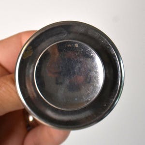 May include: A circular, metallic object with a polished, reflective silver-toned surface. The item features a smooth, slightly concave center surrounded by a raised, rounded rim. The metal surface displays minor surface scratches and reflections.