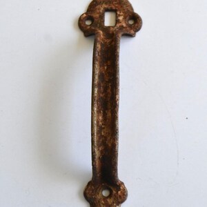 May include: A weathered, rusty brown metal door pull with a rectangular hole at the top and a rounded bottom. The handle is long and slender, with a slightly curved shape. The hardware has a vintage, antique appearance.
