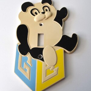 May include: A vintage panda bear light switch cover. The panda is cream-colored with black accents and is sitting on a blue and yellow block. The block has the letter "G" and "E" on it.