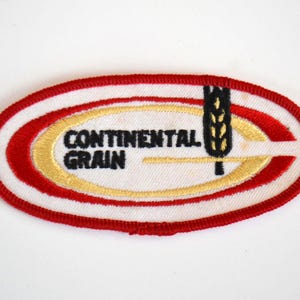 Op de afbeelding: Ovale patch met een witte achtergrond, rode rand en rode en gouden accenten. De tekst "CONTINENTAL GRAIN" is in zwart, met een gestileerde tarwe-aar grafiek. Waarschijnlijk een embleem van een bedrijf of organisatie.