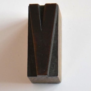 May include: A dark brown wooden letter "V" block, likely for printing or crafting. The letter is carved with a deep groove, creating a distinct shape. The block is rectangular and sits on a white surface.