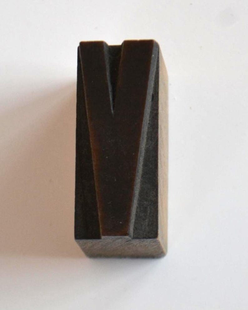 May include: A dark brown wooden letter "V" block, likely for printing or crafting. The letter is carved with a deep groove, creating a distinct shape. The block is rectangular and sits on a white surface.