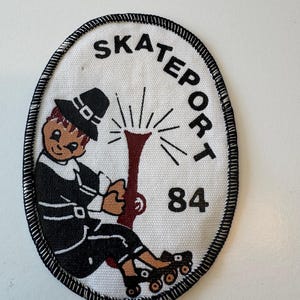 May include: Oval patch with a black border and the text "SKATEPORT" in black. The patch features a cartoon figure wearing roller skates and a hat, holding a torch. The number "84" is printed on the right side of the patch.