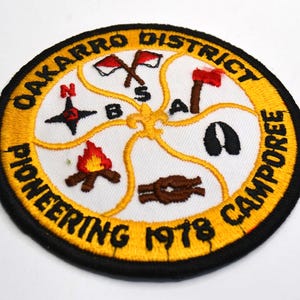 Puede incluir: Parche bordado con borde amarillo y ribete negro. El parche presenta el texto "OAKARRO DISTRICT PIONEERING CAMPOREE 1978" alrededor del borde. El centro tiene una brújula, hachas, una fogata y un nudo.