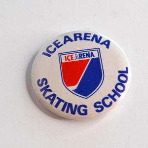 May include: A round, white button with the words "ICE ARENA SKATING SCHOOL" in blue. The center features a shield design with red and blue elements, and the text "ICE ARENA" in a rectangle.