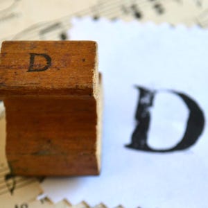 May include: A vintage wooden letterpress stamp with the letter "D" printed on its top surface. The stamp is positioned next to a printed impression of the letter "D" on a piece of paper. The background includes sheet music.