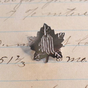 Może przedstawiać: Srebrna przypinka w kształcie liścia z ozdobną literą "M" pośrodku. Przypinka ma szczegółowe żyłkowanie i teksturowaną powierzchnię. Tło przedstawia odręczny skrypt na postarzanym papierze.