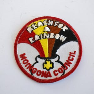 Pode incluir: Um remendo bordado em vermelho, branco e preto com o texto "Reach for a Rainbow" e "Moingona Council".
