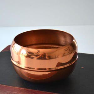 May include: A copper-coloured, round bowl with a reflective surface. The bowl has a wide opening and a slightly raised base. The interior and exterior surfaces are smooth and polished, reflecting the surrounding environment.