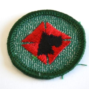 Puede incluir: Parche bordado circular con borde verde. El diseño central es un rombo rojo con centro negro. El fondo presenta un patrón texturizado blanco y verde.