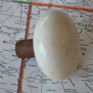 May include: A white ceramic door knob with a brown metal base. The knob is oval shaped and has a slightly textured surface.