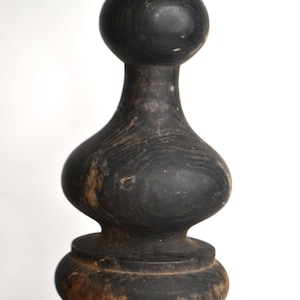 May include: A dark charcoal-colored wooden finial featuring a smooth, rounded top sphere, a tapered neck, and a bulbous midsection that rests upon a wide, circular base. The wood grain is visible throughout the surface, showing natural variations.