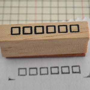 May include: A rectangular wooden rubber stamp featuring a row of six black square outlines on its top surface. The stamp is positioned above a white paper surface showing a stamped impression of the same six black square outlines in a horizontal row.