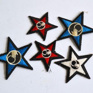 Pode incluir: Cinco patches bordados para passar a ferro em forma de estrelas. Cada estrela apresenta uma combinação de cores diferente de vermelho, branco e azul. O centro de cada estrela tem um círculo preto com uma mão branca no seu interior.